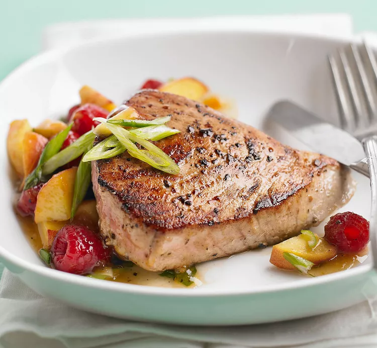 A cooked tuna steak with veggies and fruits beautifully presented on a white plate